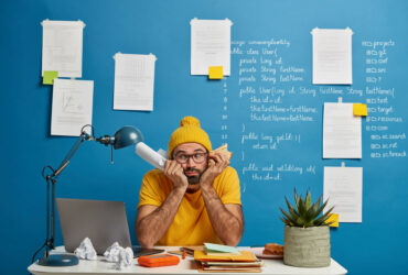 Sad male student preparess to write exam testing, poses at coworking space, holds paper and burger, wears yellow clothes, sits at dekstop with laptop, poses at modern workplace over blue wall